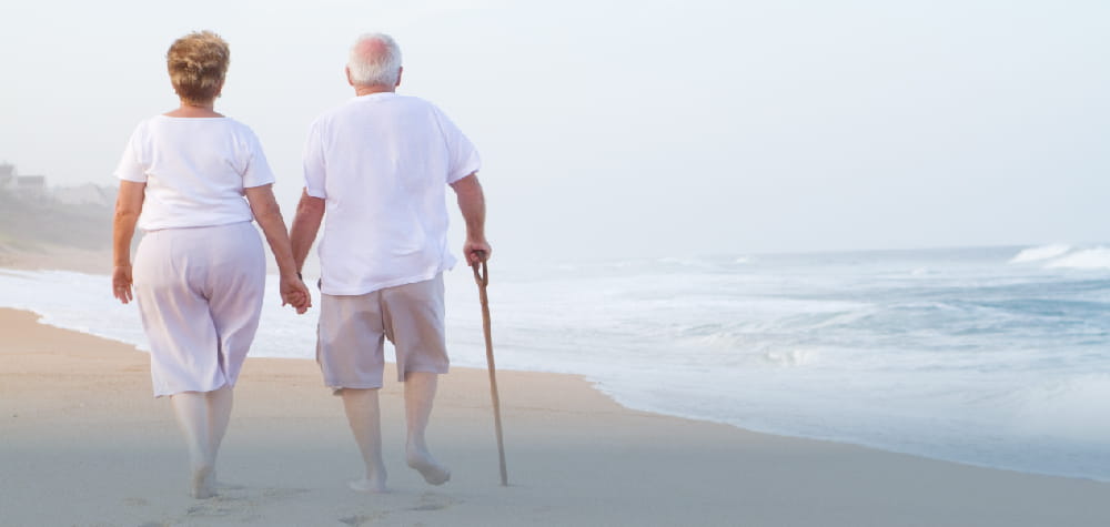 elderly retired white couple in white aging in place walking on beach barefoot
